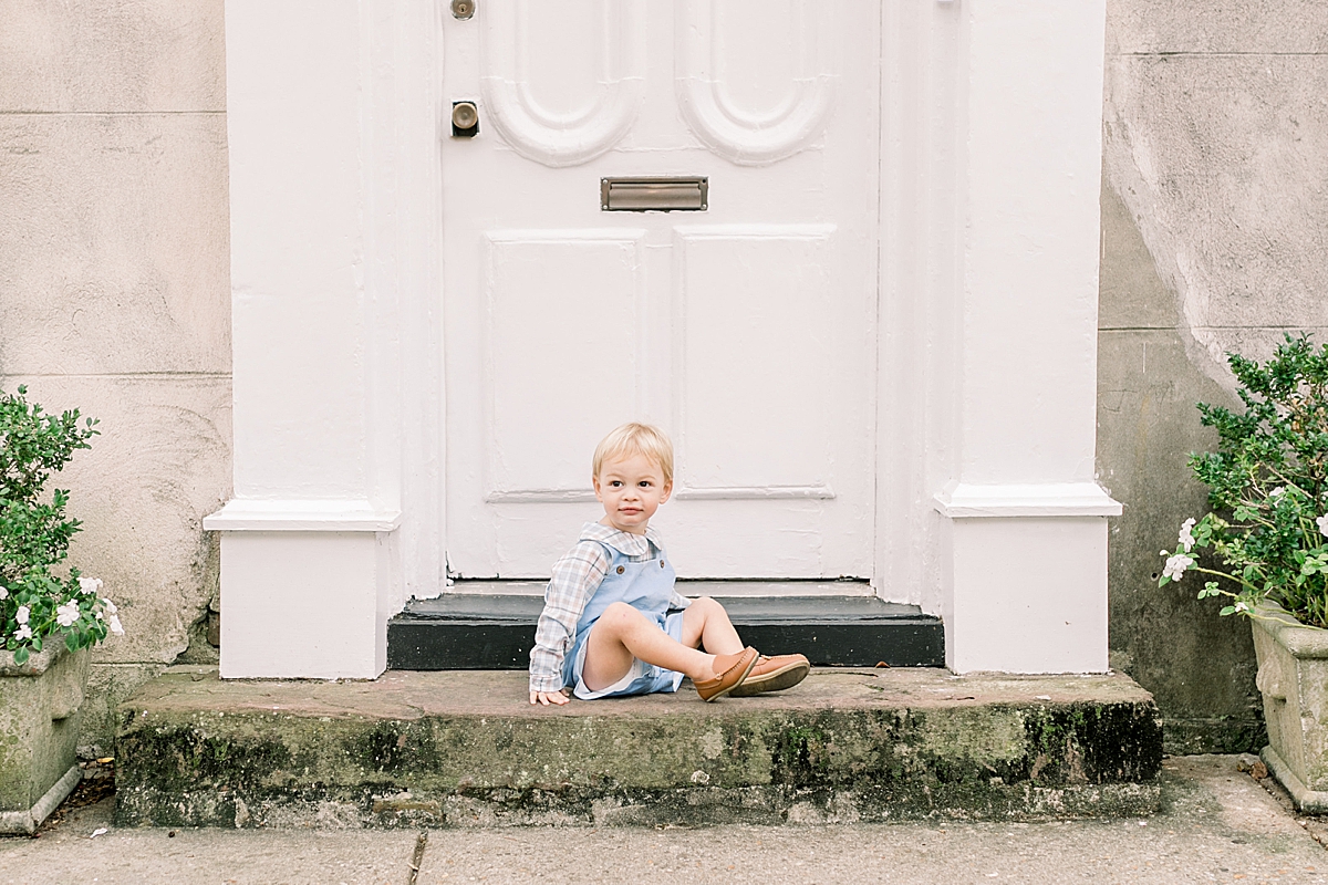 Young toddler boy sitting on a front porch in Downtown Charleston. Family photographer in Charleston photographs family downtown on the cobblestone streets. Photos by Caitlyn Motycka Photography.