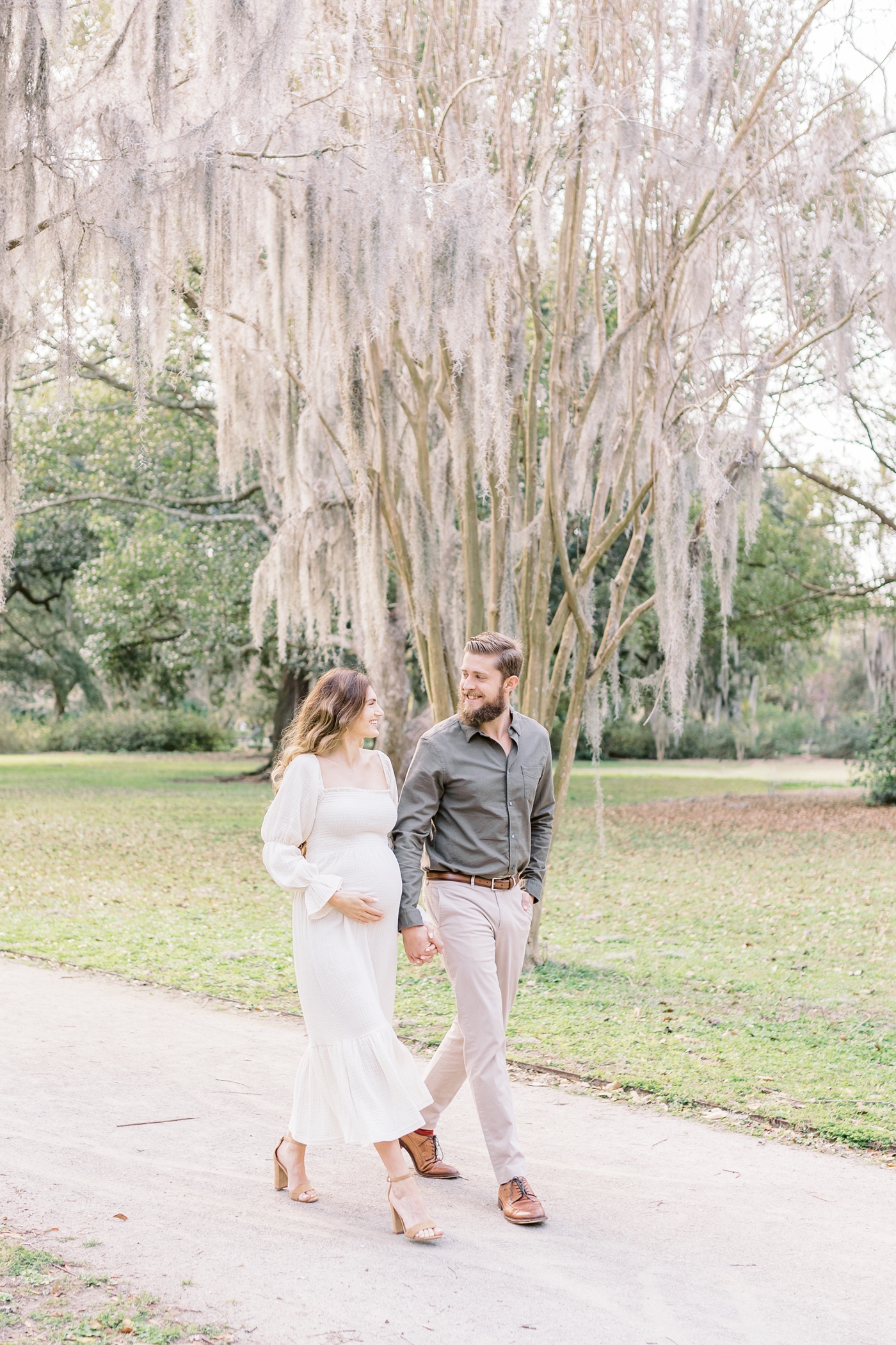 Expecting parents smiling at each other while walking on path in park during session with Charleston photographer, Caitlyn Motycka Photography.