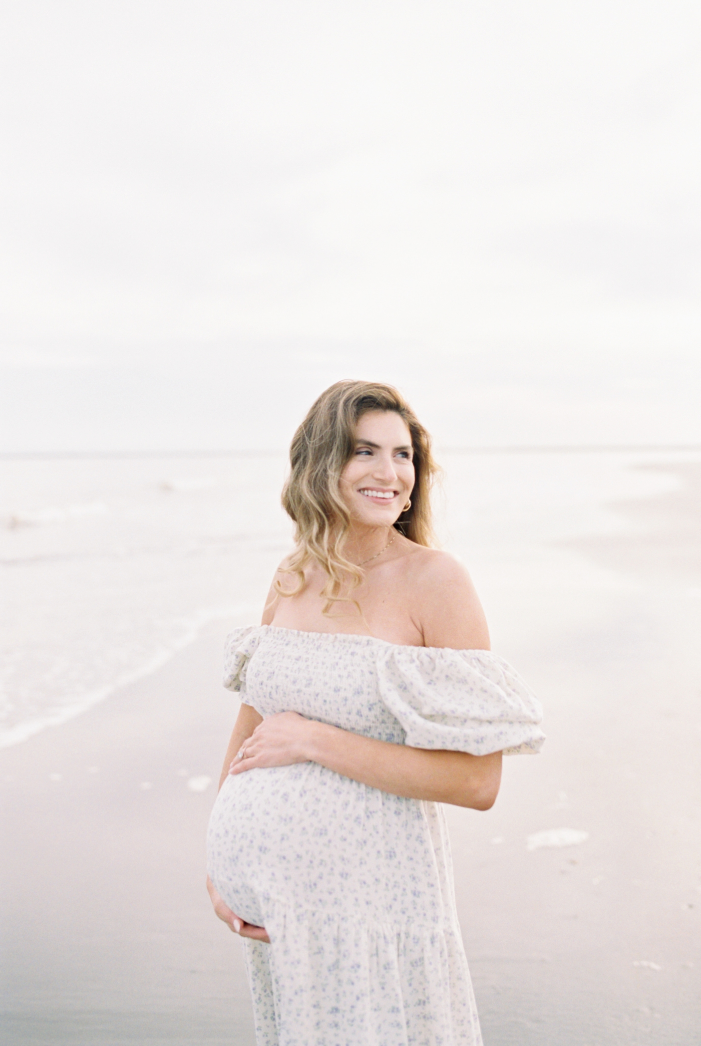 Mom to be in white dress with blue florals on the beach | Photo by Caitlyn Motycka Photography.