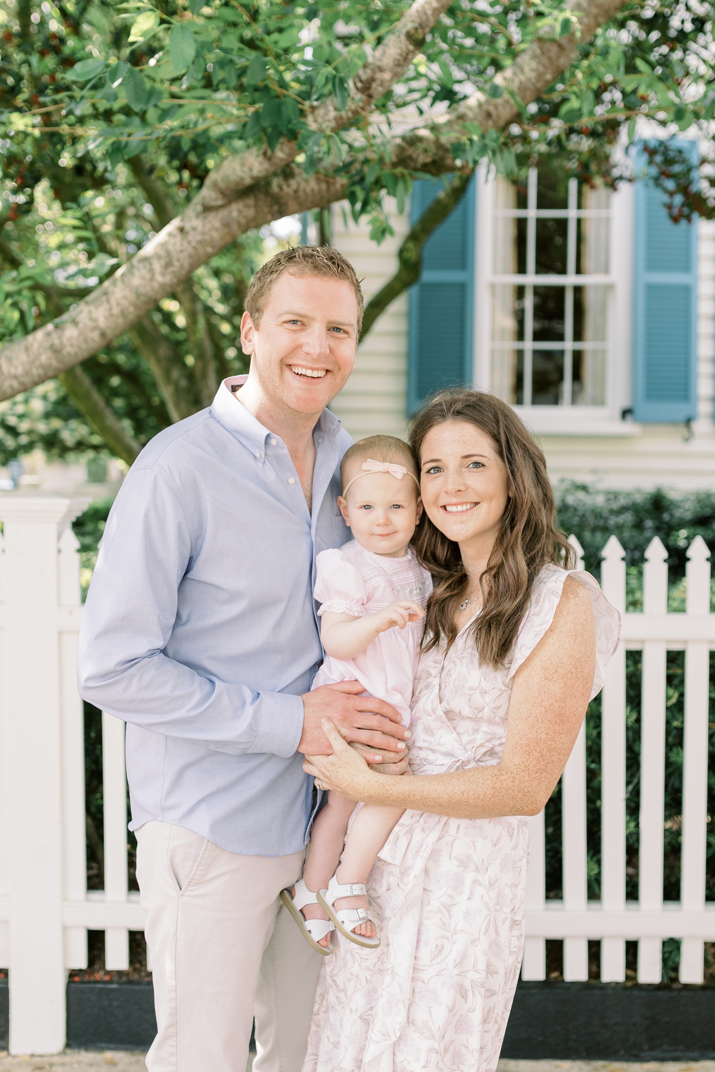 Mom and dad with baby girl smiling in front of picket fence | Photo by Caitlyn Motycka Photography.