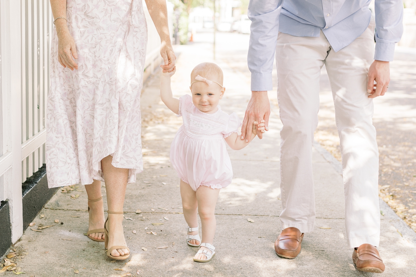 Mom and dad helping baby daughter walk down the sidewalk | Photo by Caitlyn Motycka Photography.