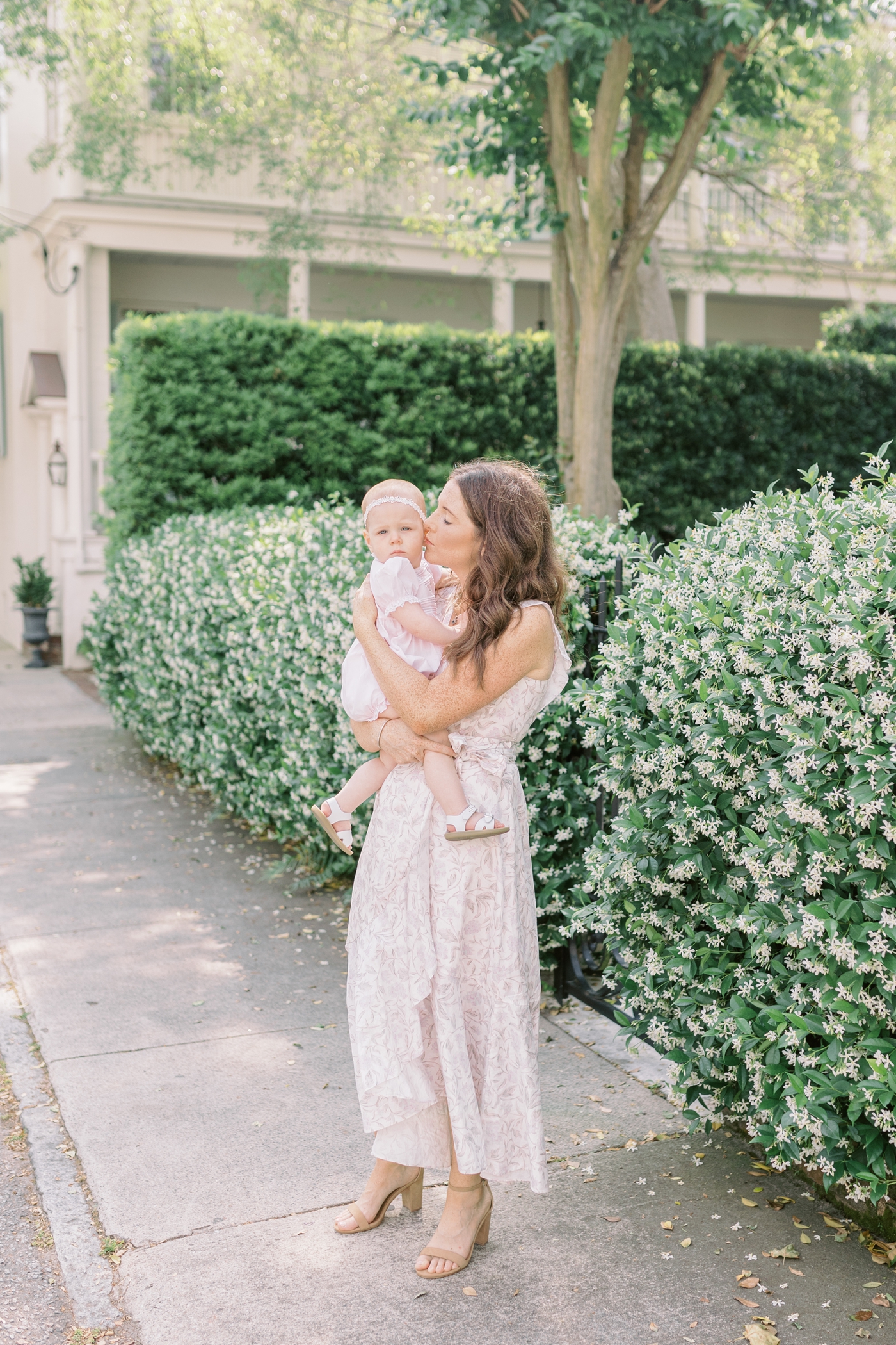 Mom kissing baby daughters cheek in front of jasmine covered sidewalk downtown charleston | Photo by Caitlyn Motycka Photography.
