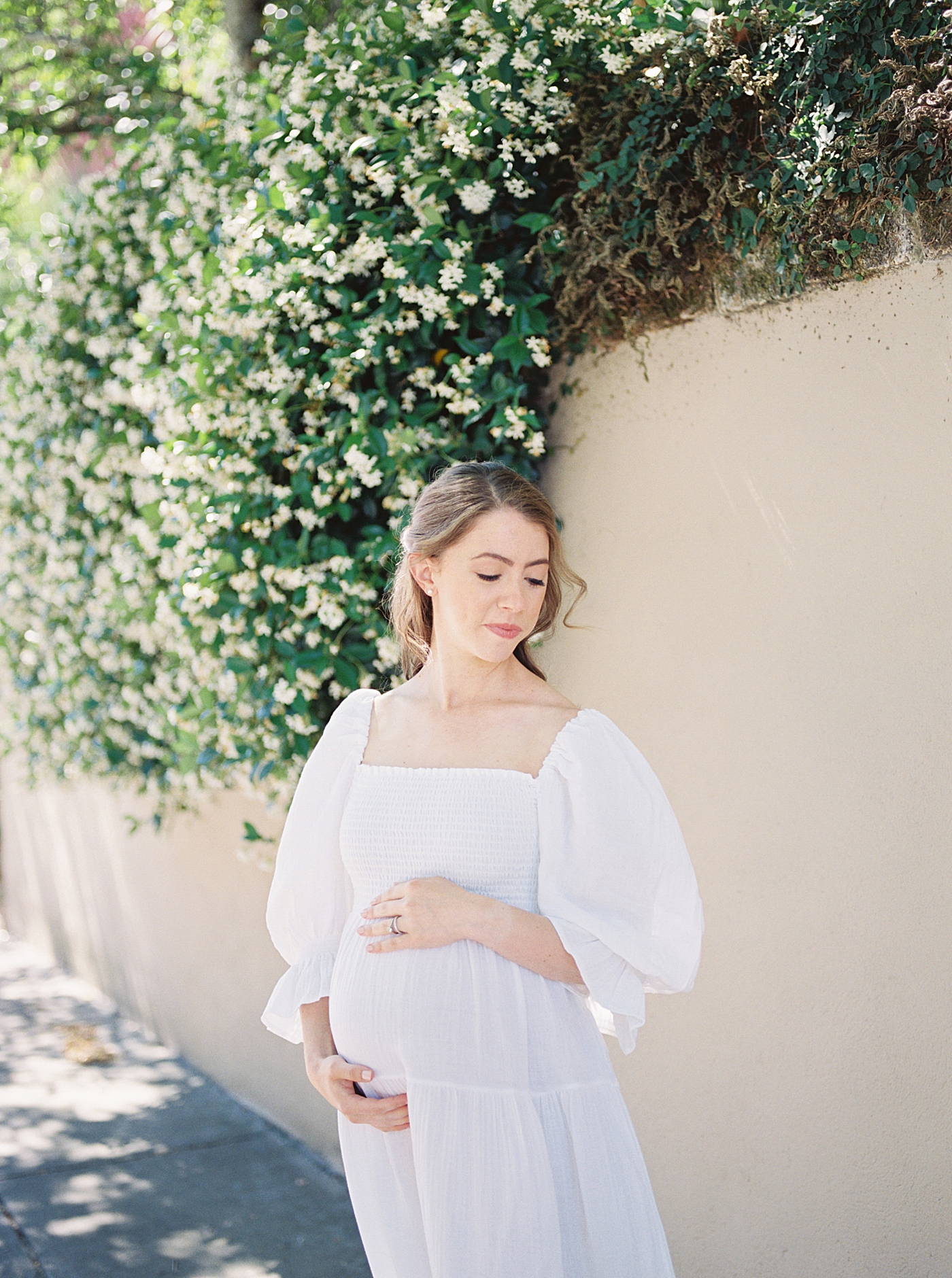 Mother to be holding her belly in front of jasmine covered wall | Photo by Charleston Film Photographer Caitlyn Motycka