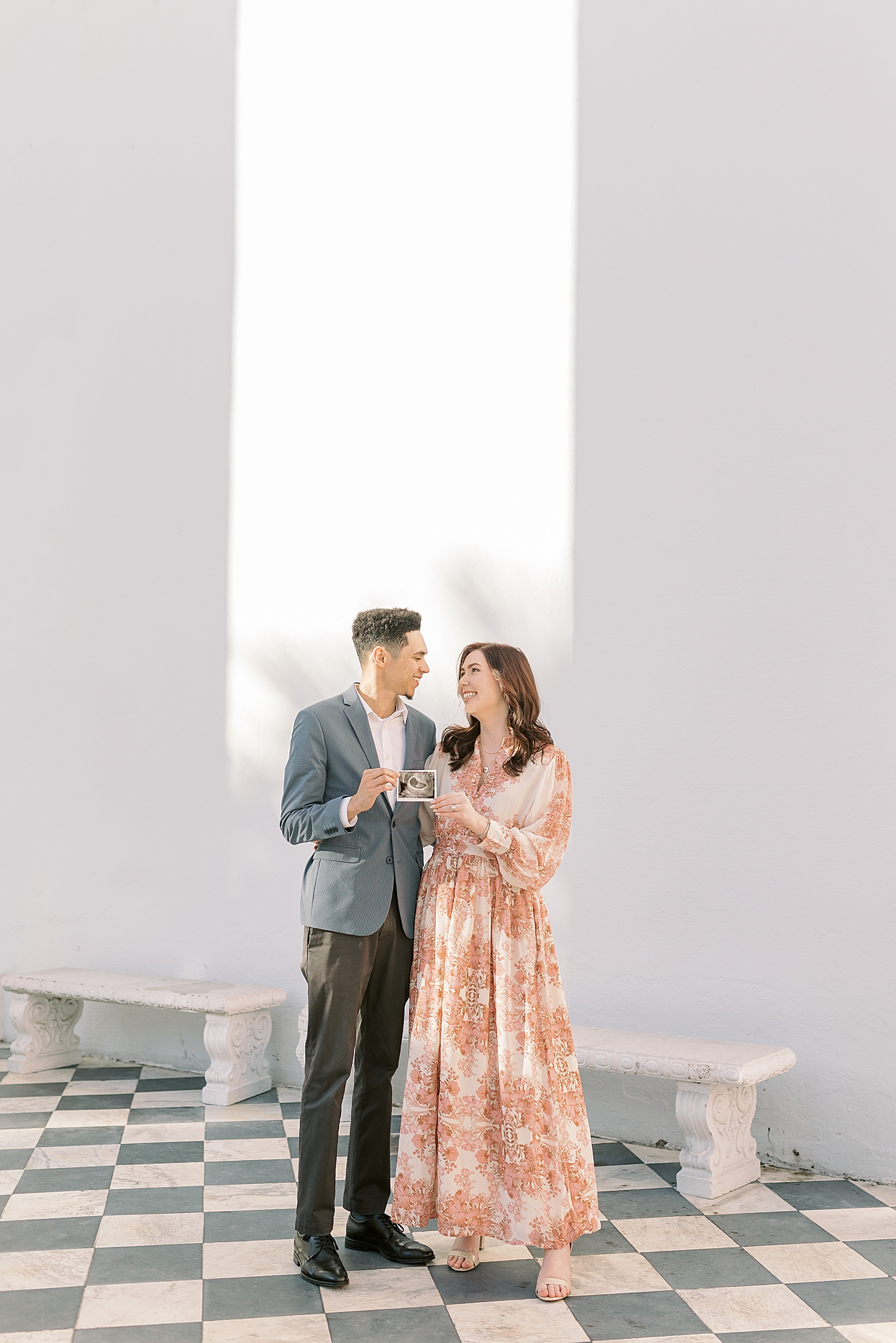 Mom and dad to be standing on checkered floor smiling at each other | Photo by Caitlyn Motycka Photography