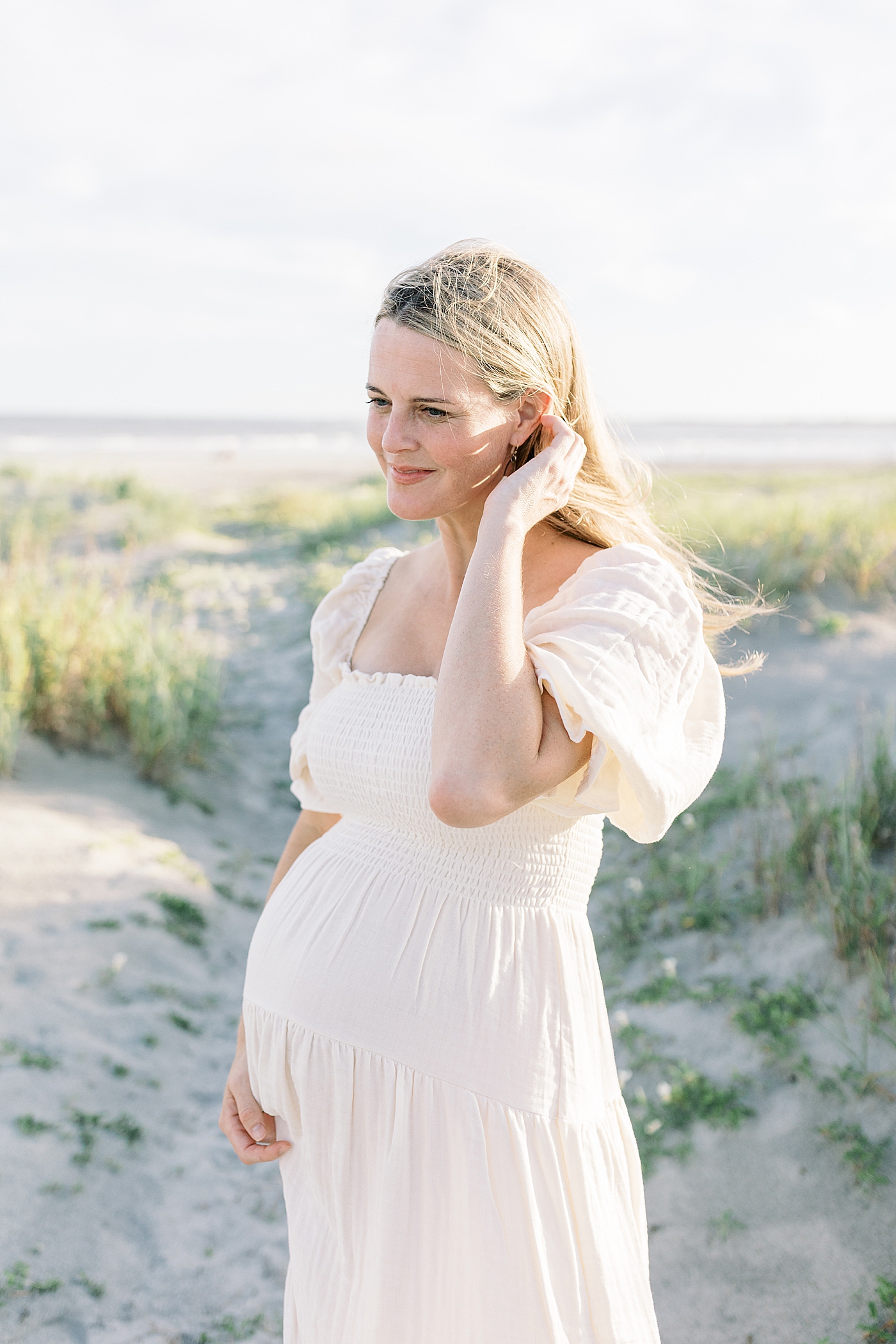 Pregnant mom in a cream dress holding her belly during Maternity Session at Folly Beach | Image by Caitlyn Motycka