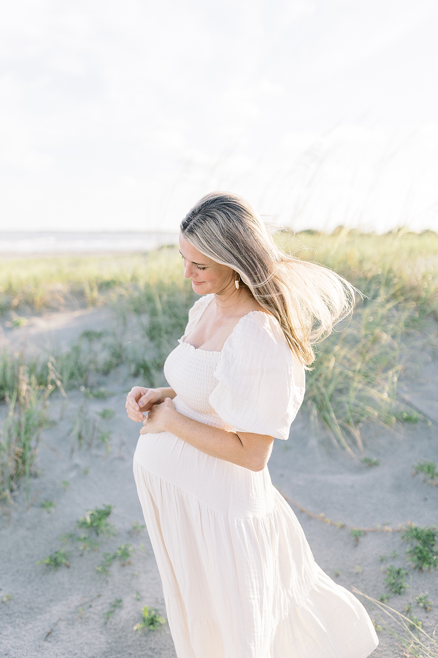 Mom to be with blonde hair in a white dress holding her belly | Image by Caitlyn Motycka