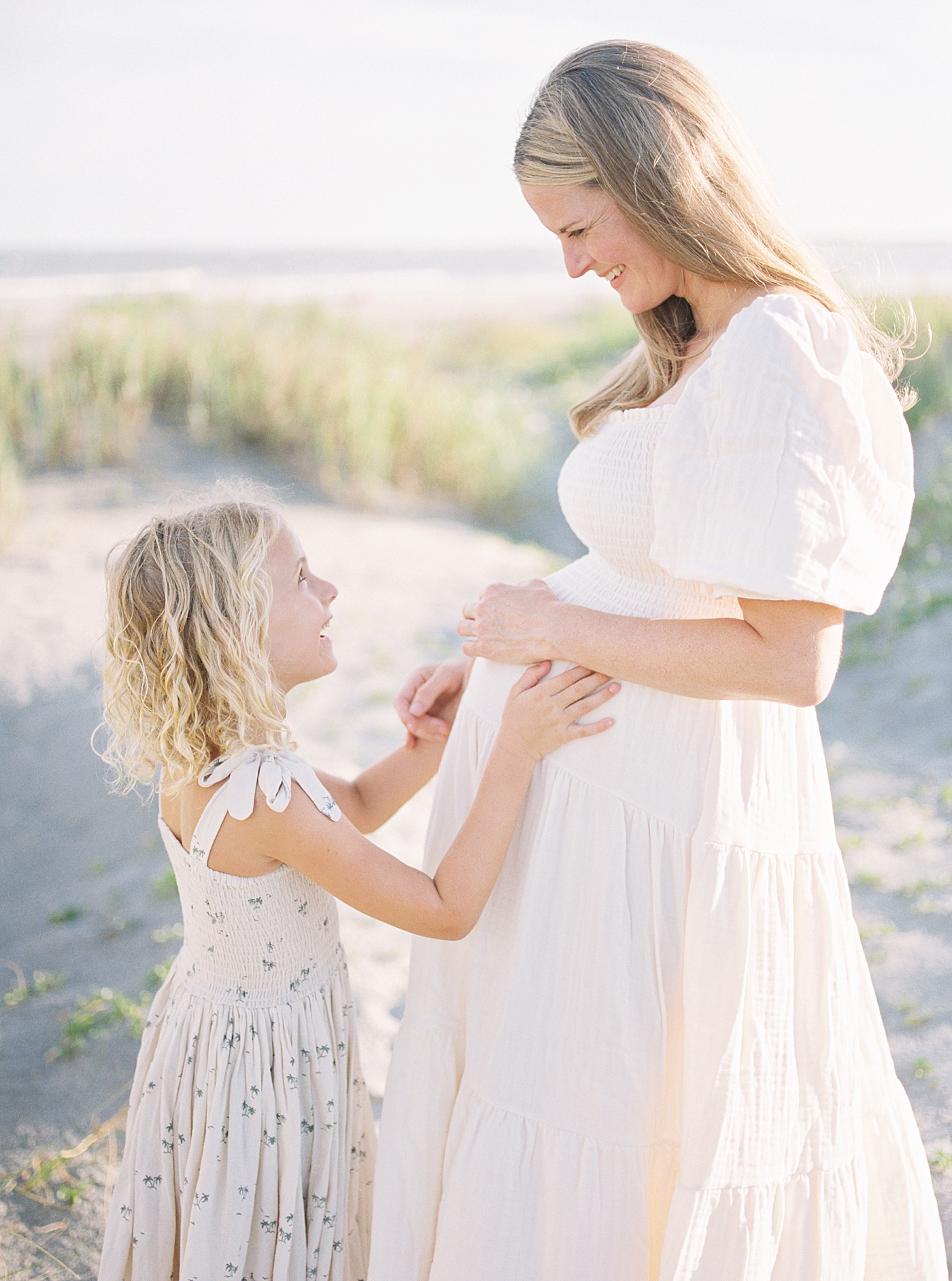 Little girl in floral dress with her hands on mom's pregnant belly | Image by Caitlyn Motycka