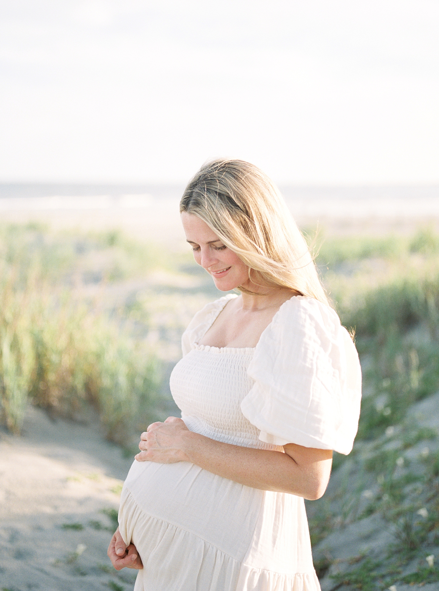 Mom in a flowy white dress with her hand resting on her pregnant belly | Image by Caitlyn Motycka