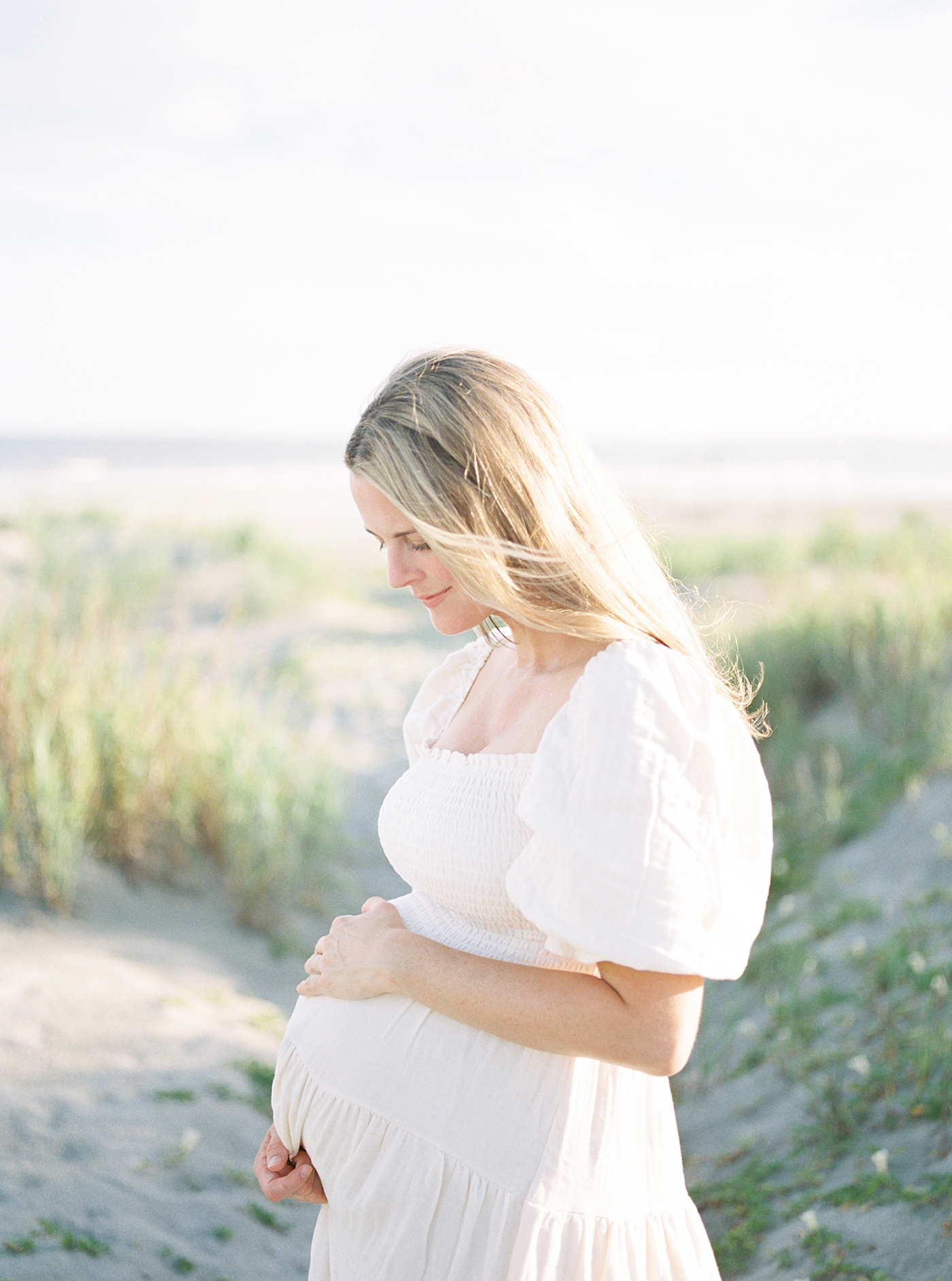 Mom with long blonde hair in a white dress holding her belly on the beach | Image by Caitlyn Motycka