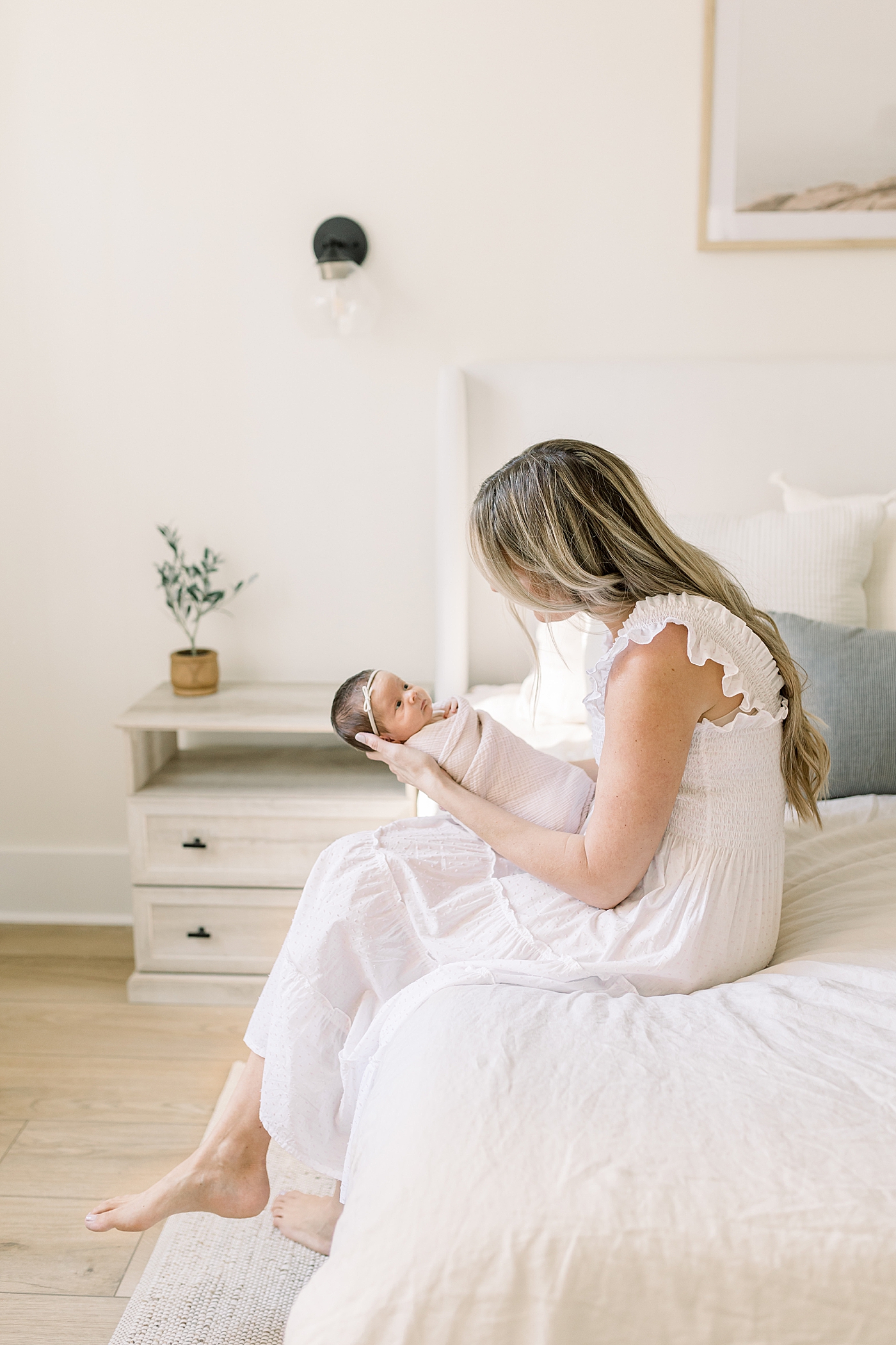 Mom in white dress sitting on the edge of her bed holding newborn baby girl | Image by Caitlyn Motycka