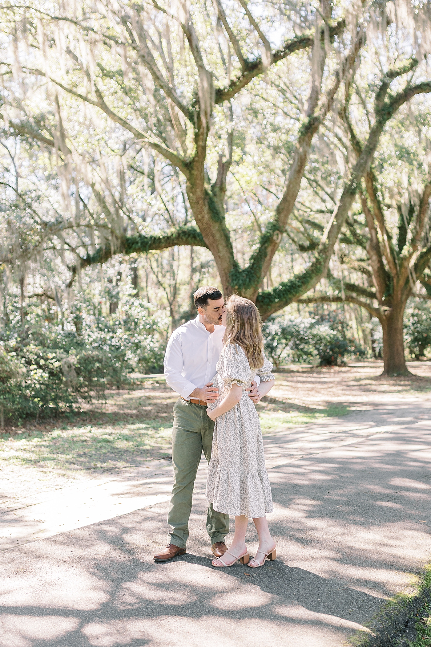 Husband and wife kissing and holding her pregnant belly on an oak tree-lined walkway Maternity Session at Charlestowne Landing| Image by Caitlyn Motycka Photography