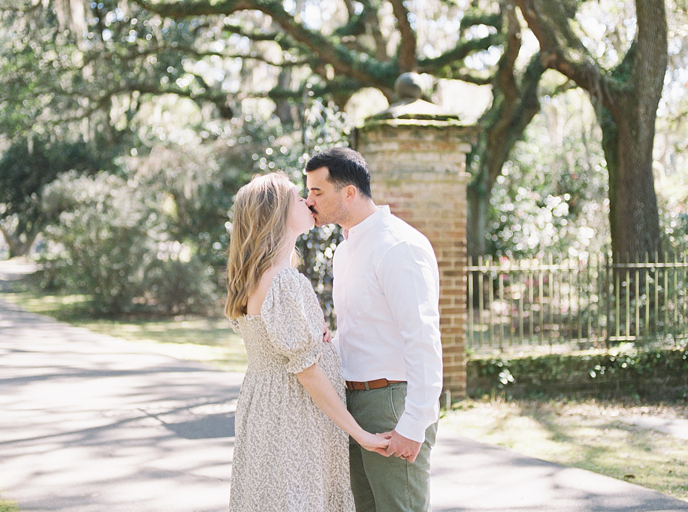 Husband and an expecting wife in a spring dress kissing and holding hands on an oak tree-lined walkway | Image by Caitlyn Motycka Photography