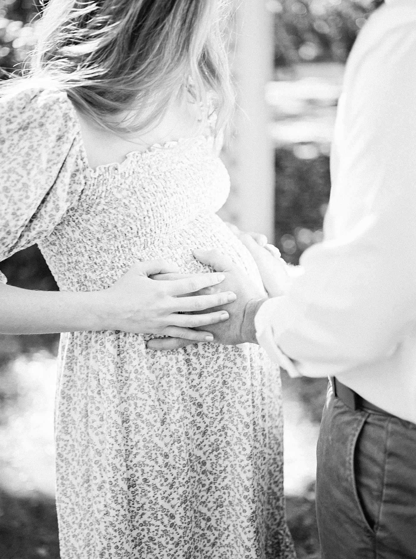 Black-and-white close up image of husband and wife in a spring dress holding her pregnant belly on an oak tree-lined walkway | Image by Caitlyn Motycka Photography