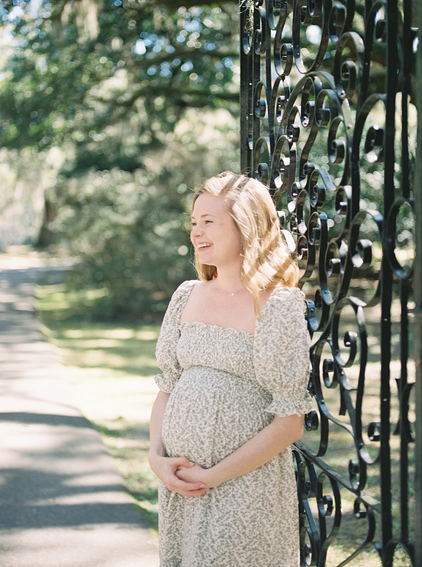 Woman in a spring dress holding her pregnant belly next to an iron gate during their Maternity Session at Charlestowne Landing| Image by Caitlyn Motycka Photography