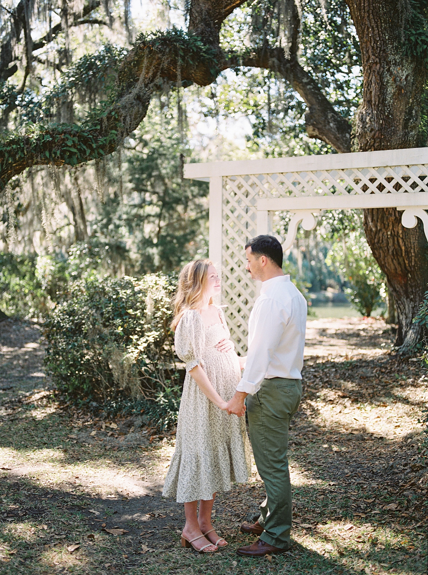 Husband and wife in a spring dress holding hands with her husband next to an arbor under moss-covered oak trees during their Maternity Session at Charlestowne Landing| Image by Caitlyn Motycka Photography