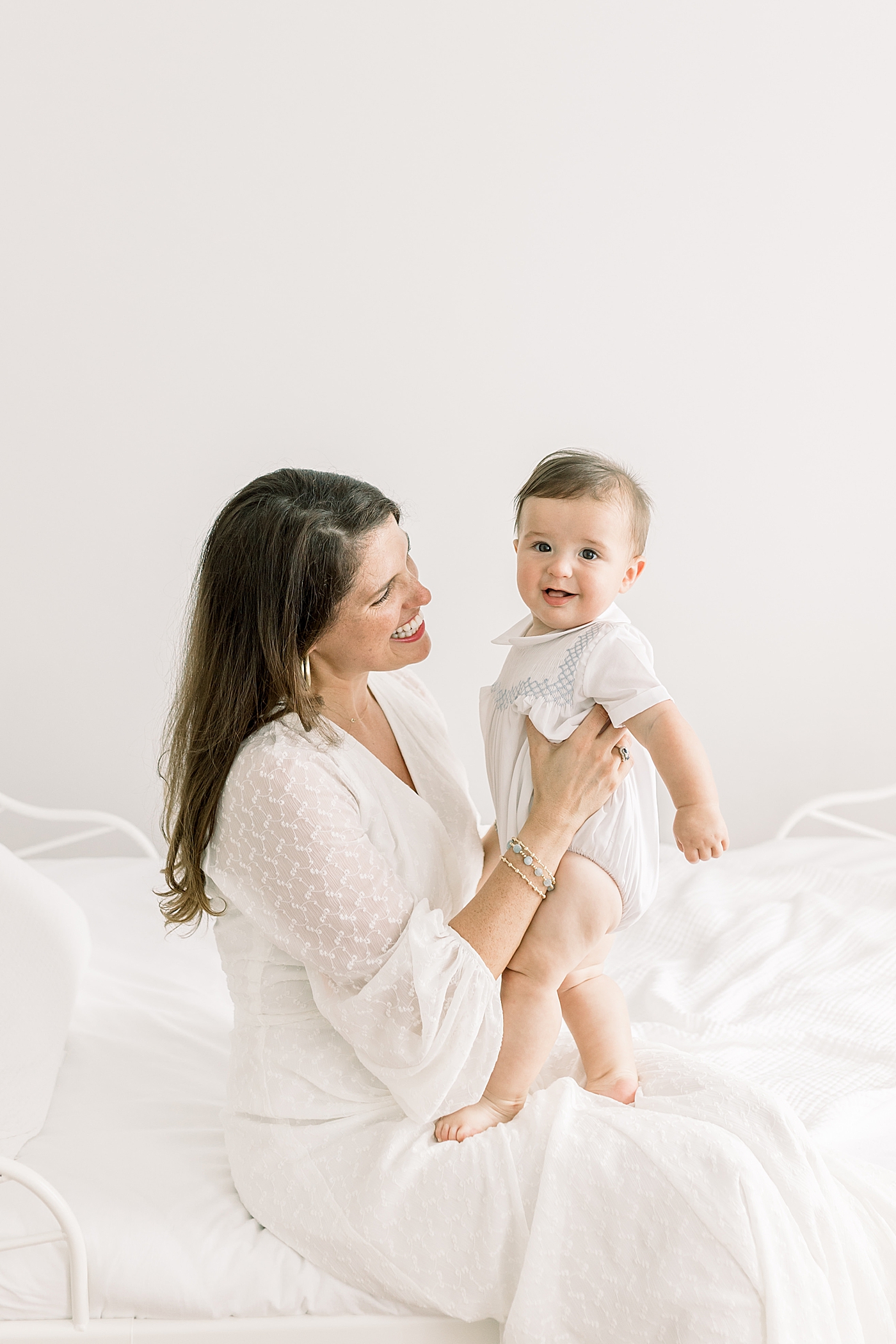 Mother looking at and holding her baby, while baby is looking at the camera in a softly lit room during Studio Milestone Session | Images by Caitlyn Motycka