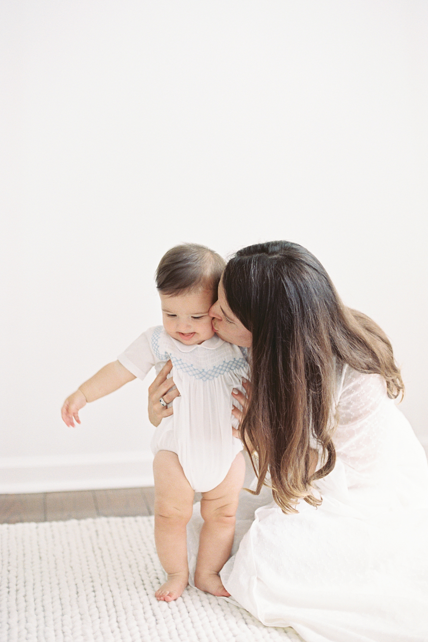 Mother kneeling, kissing and holding baby on a white room and white rug | Images by Caitlyn Motycka