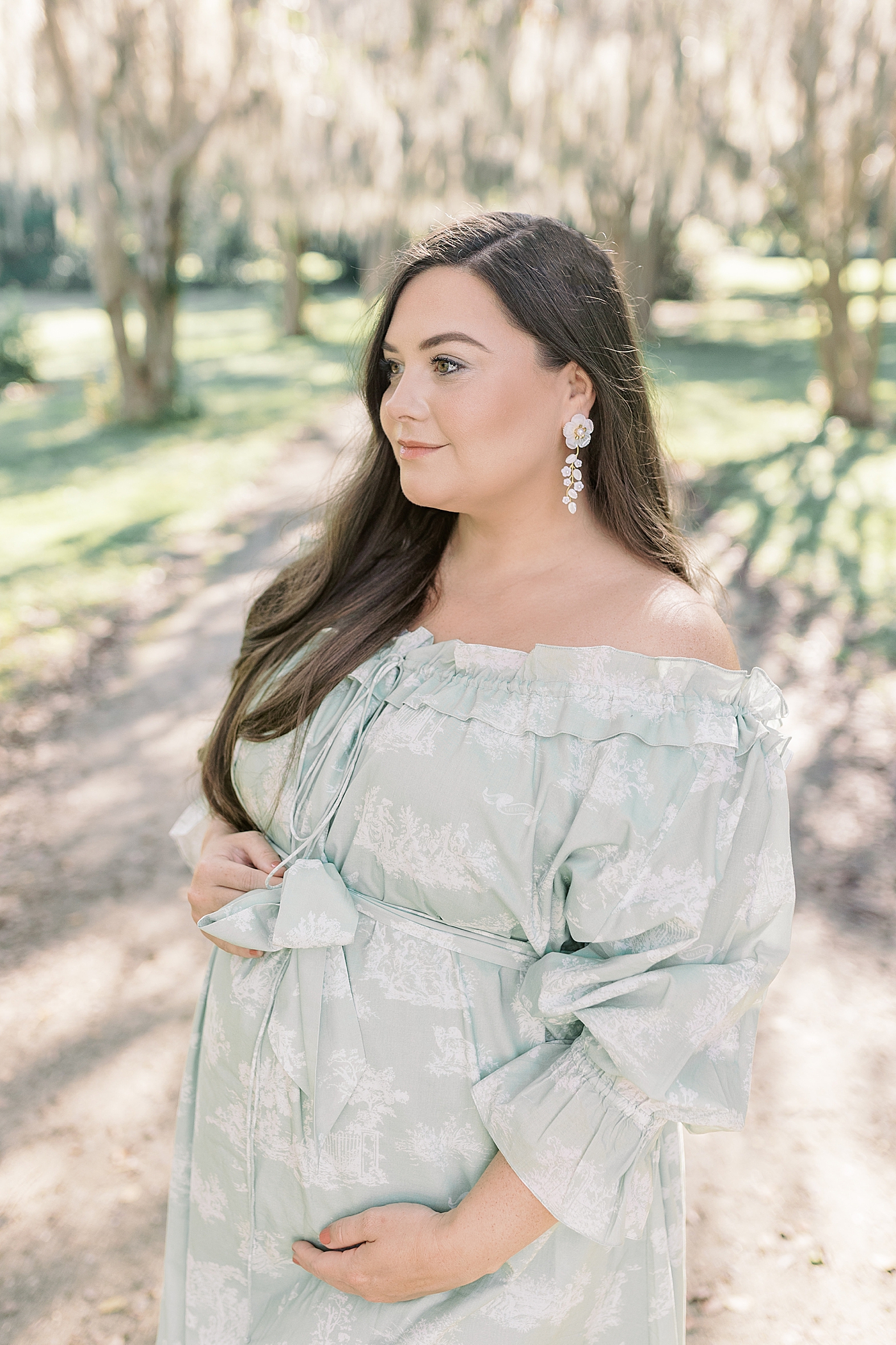 Expecting mother in a green off-the-shoulder spring dress smiling and holding her pregnant belly | Image by Caitlyn Motycka 