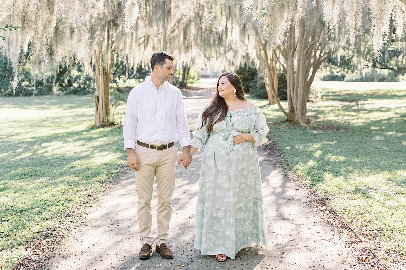 Expecting mother and husband in a green off-the-shoulder spring dress walking and looking at each other smiling, holding her pregnant belly on a tree-lined path during Spring Babymoon in Charleston | Image by Caitlyn Motycka 