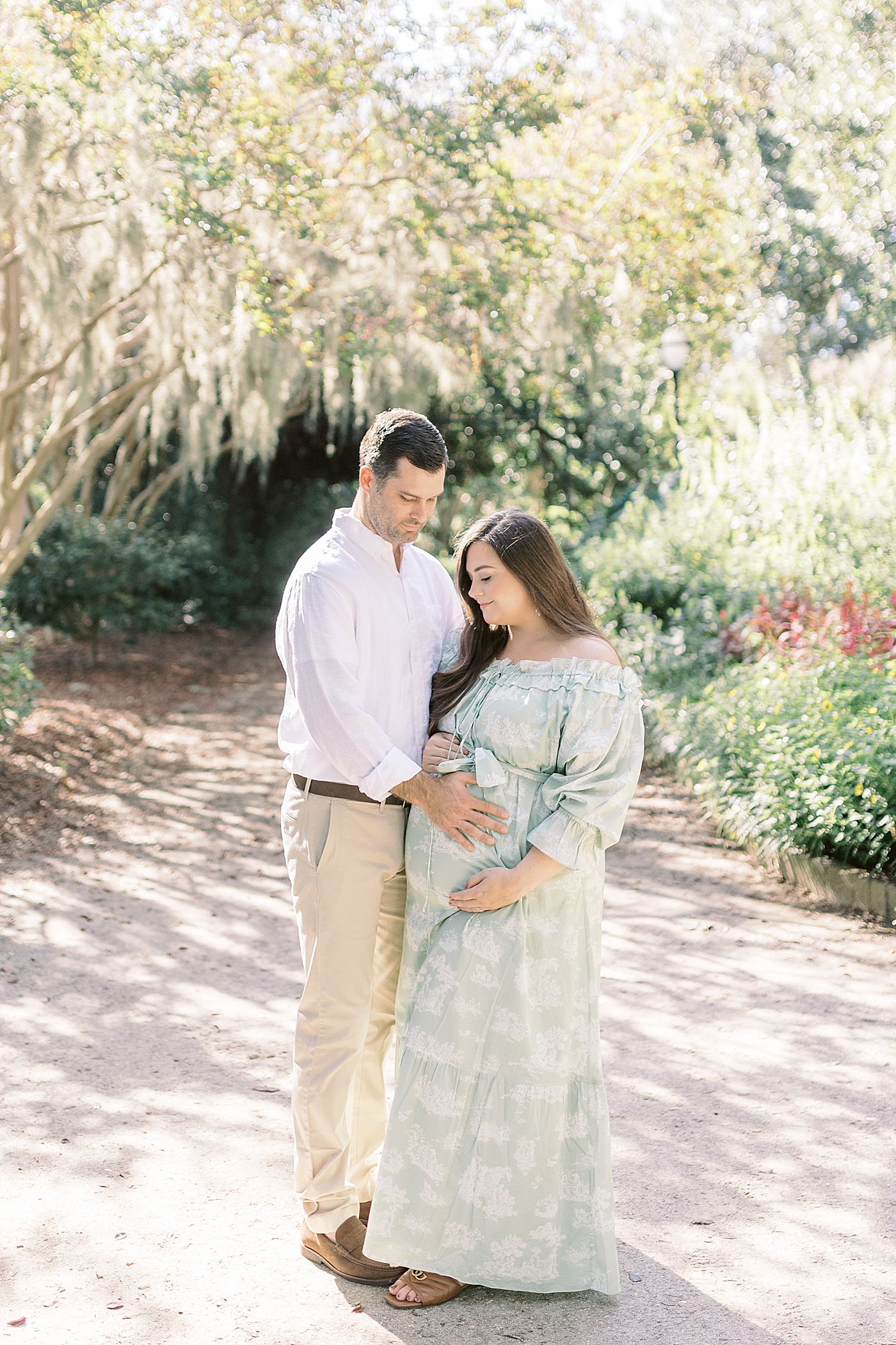 Expecting mother and husband in a green off-the-shoulder spring dress holding her pregnant belly on a tree-lined path during Spring Babymoon in Charleston | Image by Caitlyn Motycka 