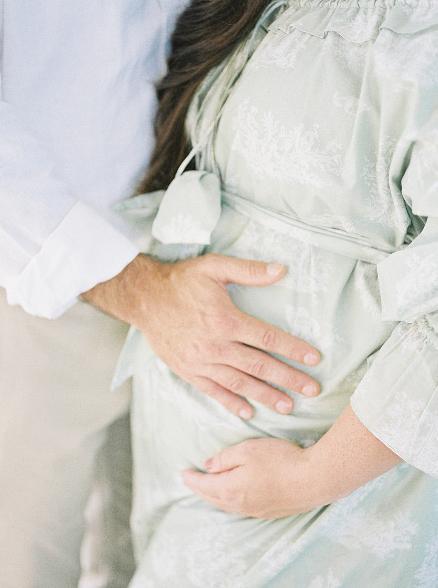Close up of expecting mother and husband in a green off-the-shoulder spring dress holding her pregnant belly | Image by Caitlyn Motycka 