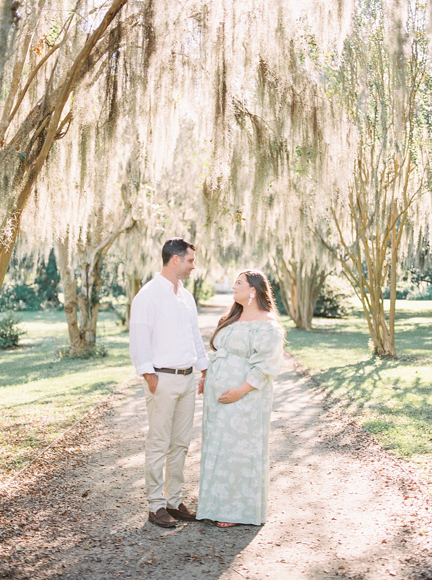 Expecting mother and husband in a green off-the-shoulder spring dress looking at each other while she is holding her pregnant belly on a tree-lined path during Spring Babymoon in Charleston | Image by Caitlyn Motycka 
