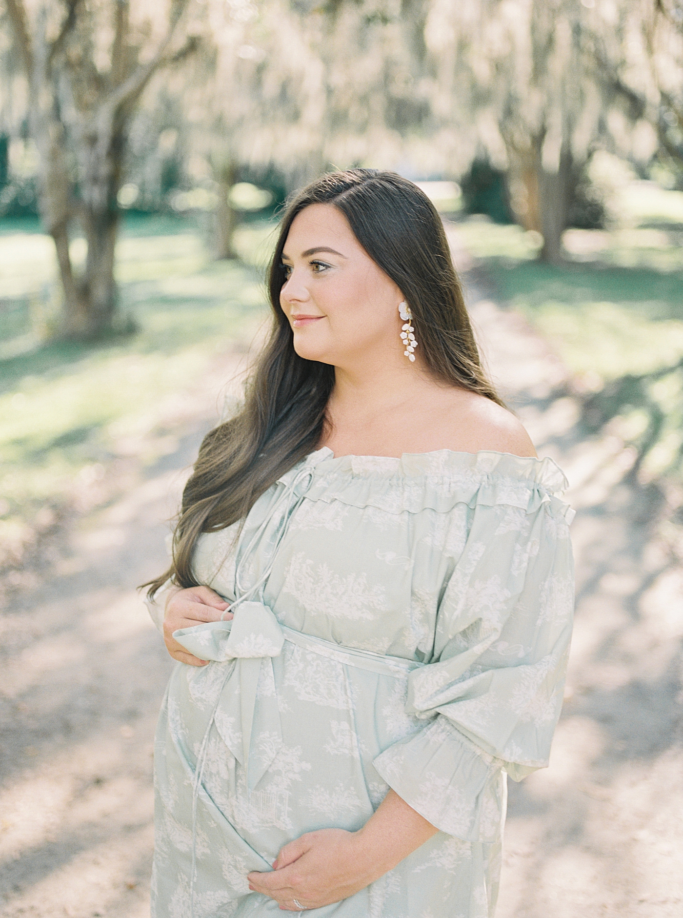 Expecting mother in a green off-the-shoulder spring dress smiling and holding her pregnant belly Spring Babymoon in Charleston | Image by Caitlyn Motycka 