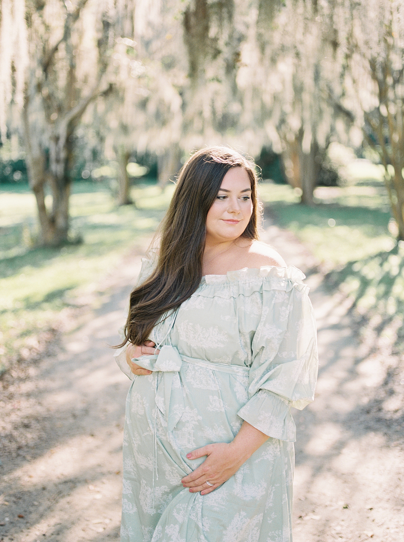 Expecting mother in a green off-the-shoulder spring dress smiling and holding her pregnant belly under oak tree-lined pathway | Image by Caitlyn Motycka 