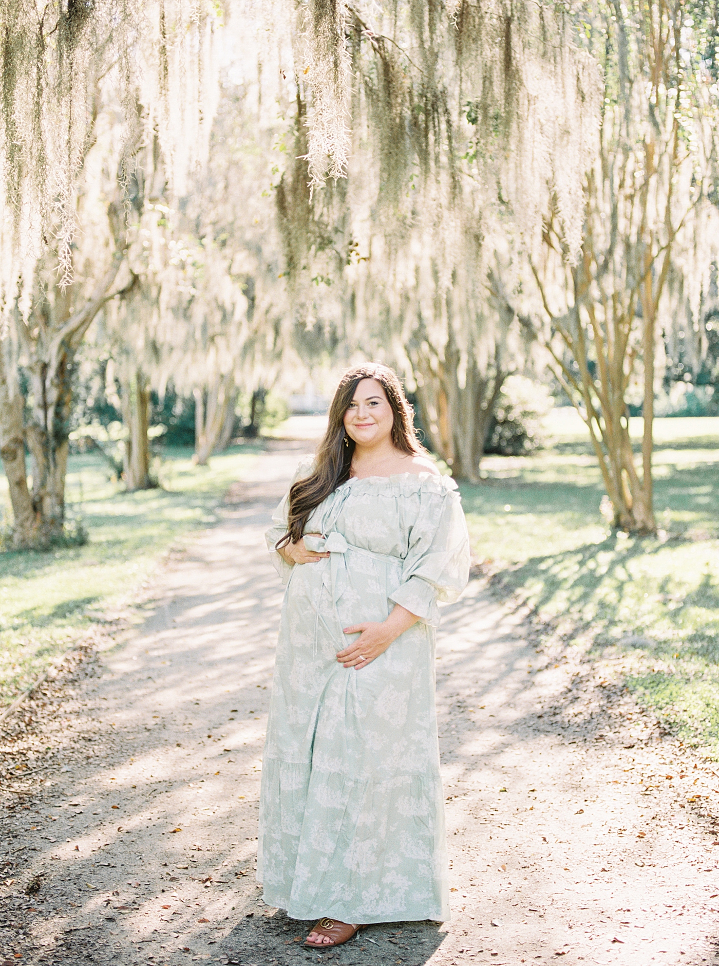 Expecting mother in a green off-the-shoulder spring dress smiling, looking at the camera, and holding her pregnant belly under oak tree-lined pathway during Spring Babymoon in Charleston | Image by Caitlyn Motycka 