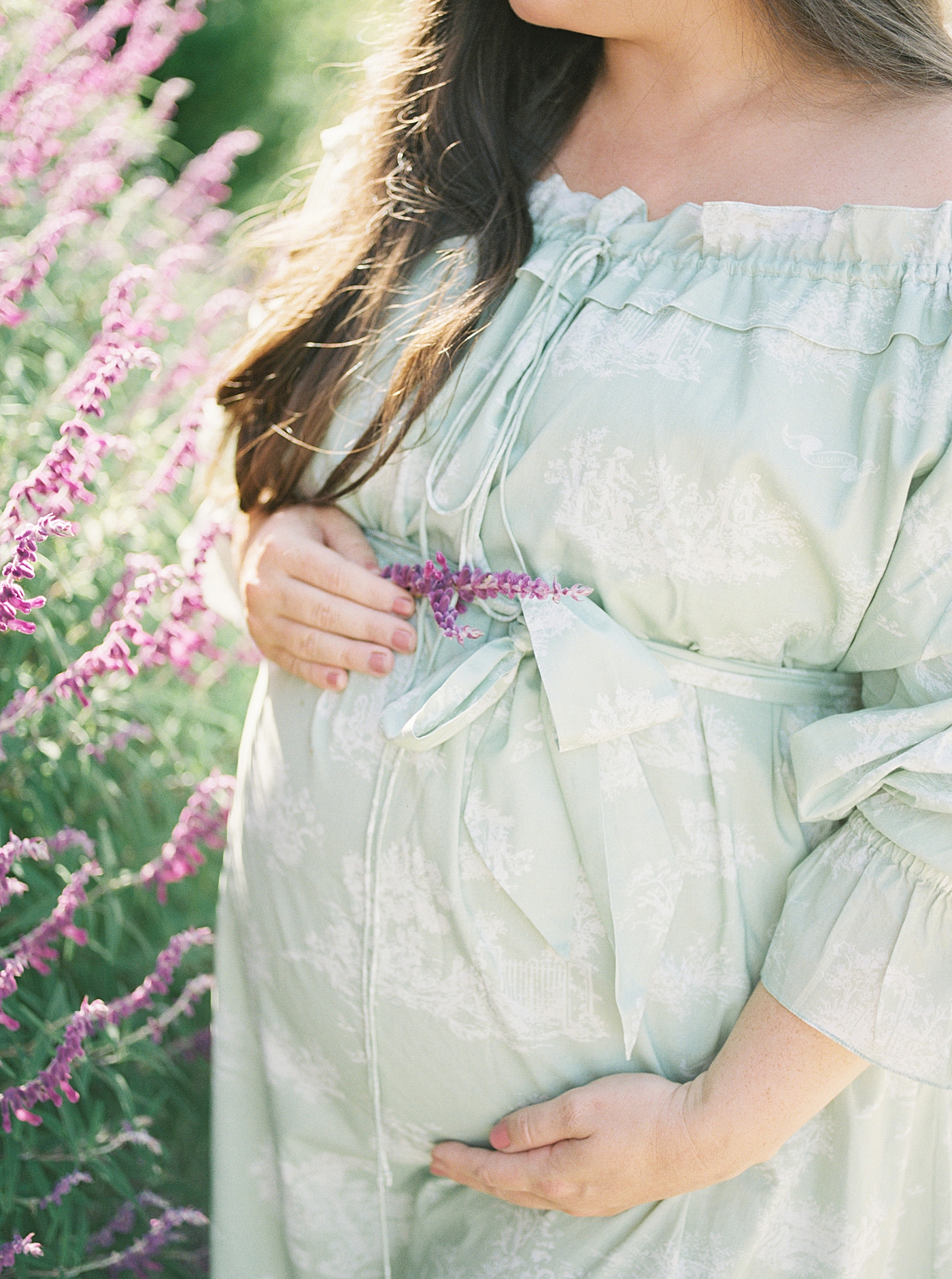 Expecting mother in a green off-the-shoulder spring dress holding her pregnant belly next to purple flowers during Spring Babymoon in Charleston | Image by Caitlyn Motycka 