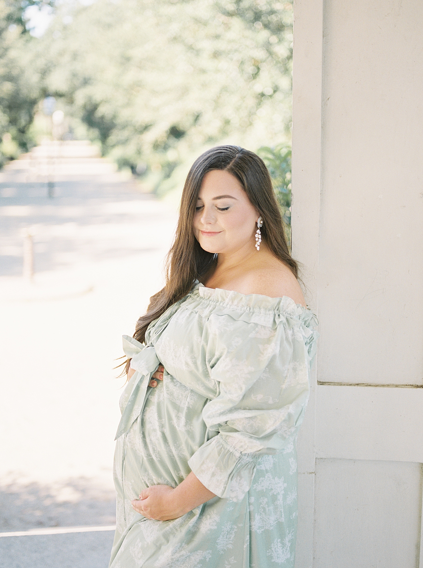 Expecting mother in a green off-the-shoulder spring dress holding her pregnant belly during Spring Babymoon in Charleston | Image by Caitlyn Motycka 