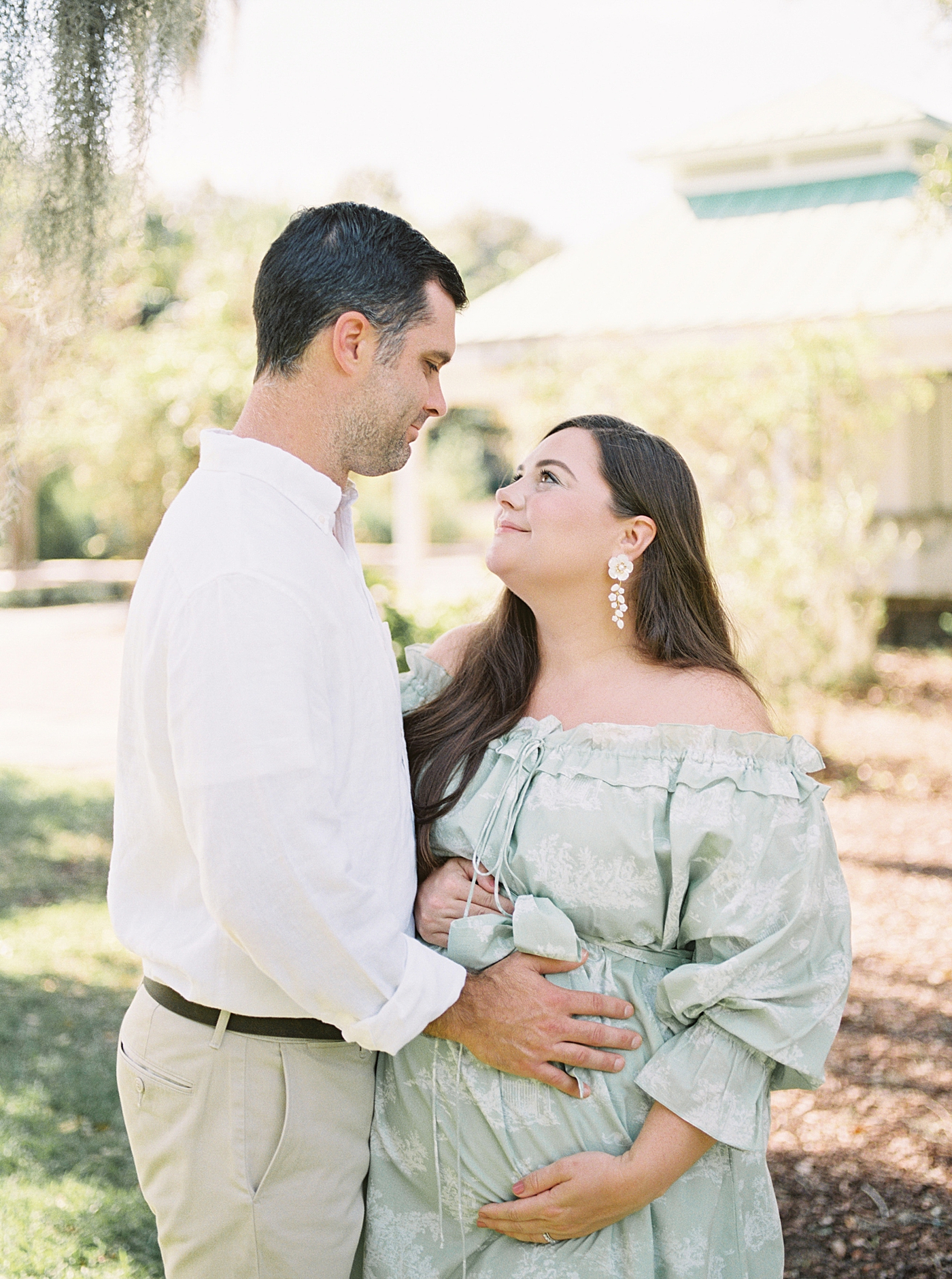 Expecting mother and husband in a green off-the-shoulder spring dress, looking at each other while she holds her pregnant belly during Spring Babymoon in Charleston | Image by Caitlyn Motycka 