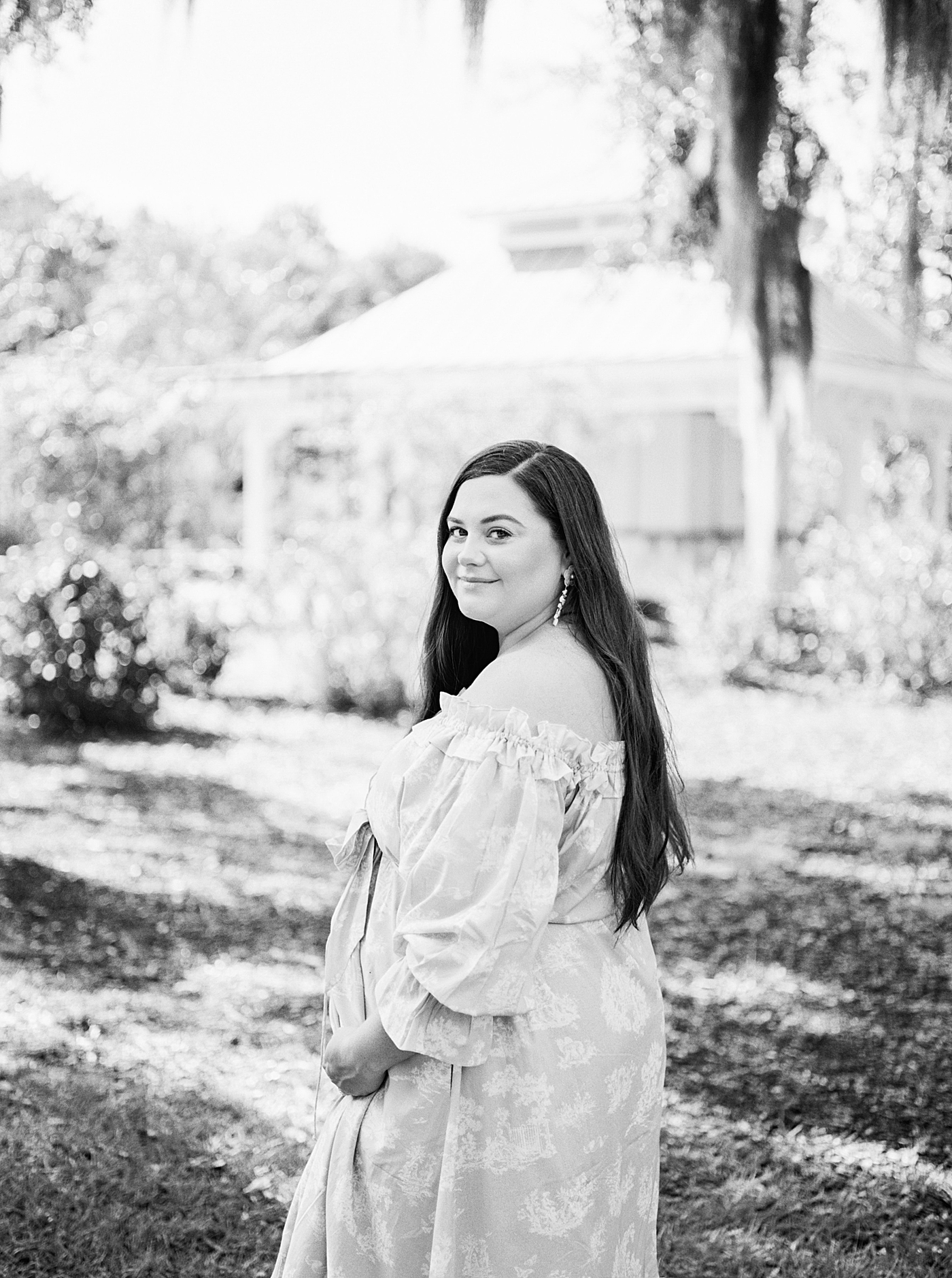 Black and white image of expecting mother in a green off-the-shoulder spring dress holding her pregnant belly during Spring Babymoon in Charleston | Image by Caitlyn Motycka 