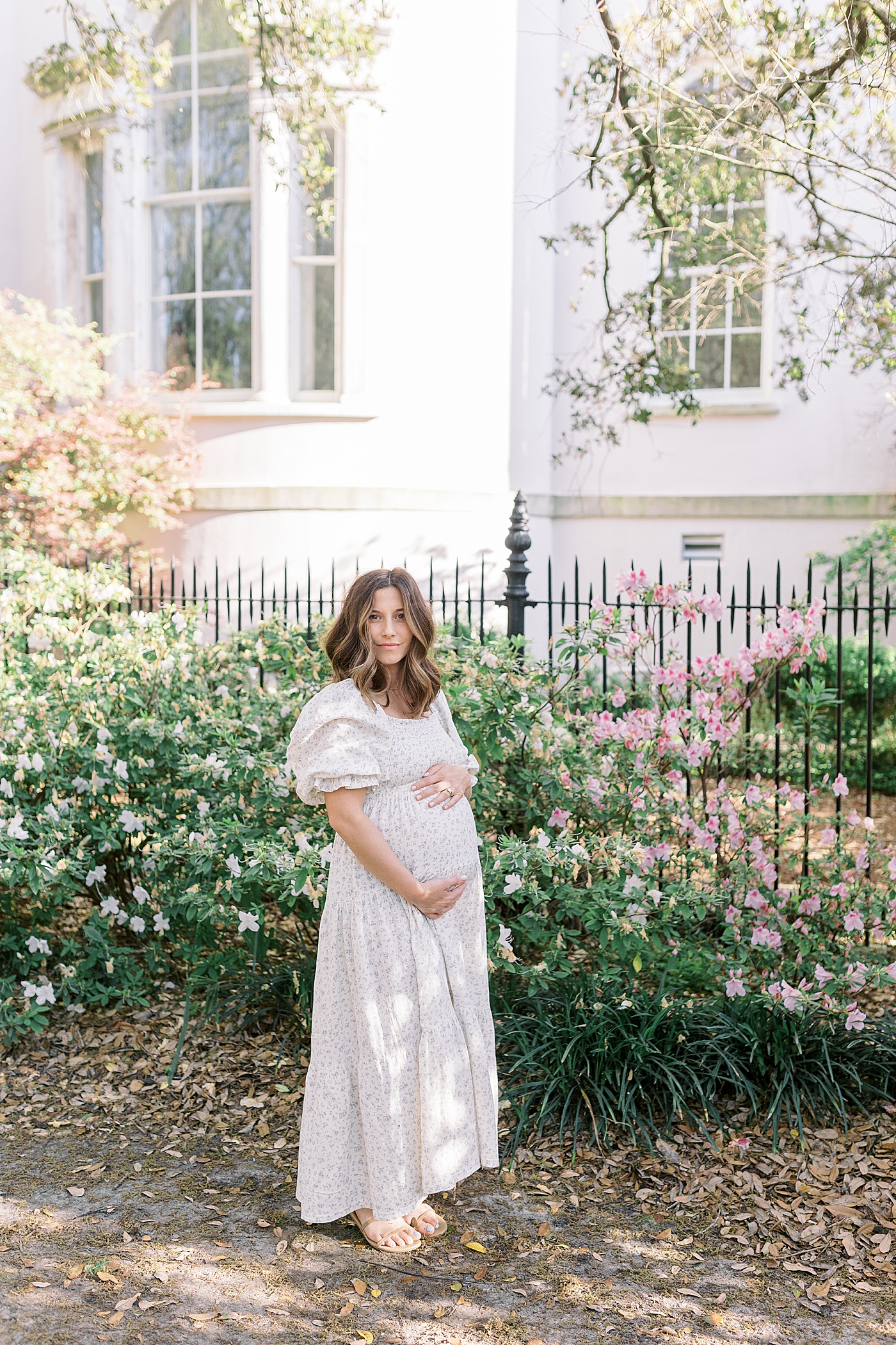 Expecting mother standing beside a home with flowers and a iron gate holding her stomach during Spring Motherhood Minis in Charleston | Image by Caitlyn Motycka