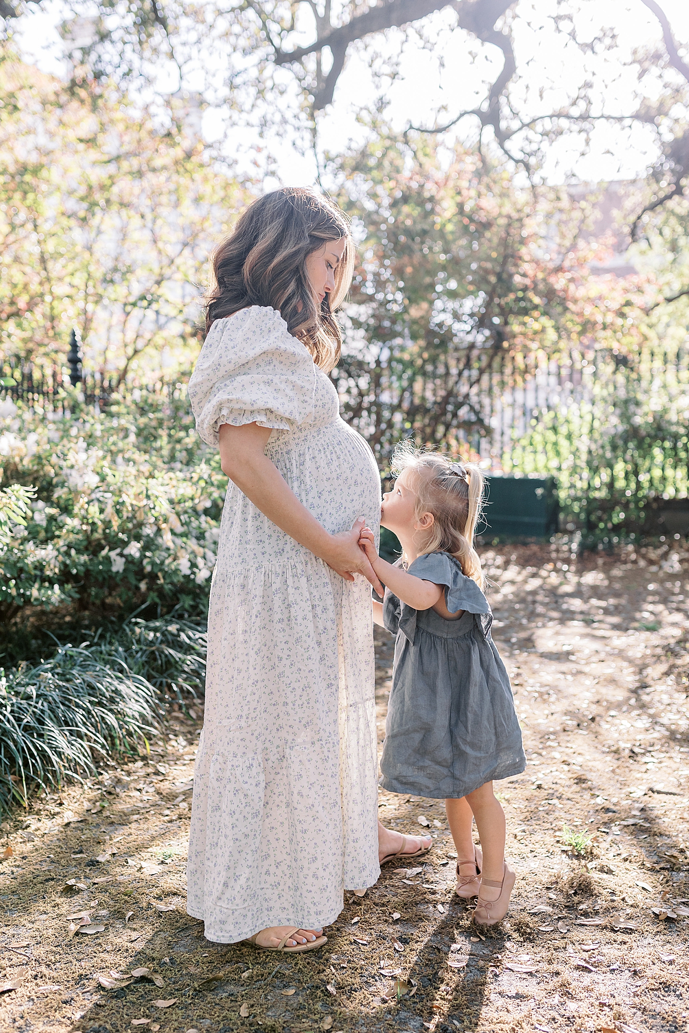 Expecting mother standing with daughter kissing her stomach in a garden with an iron gate during Spring Motherhood Minis in Charleston | Image by Caitlyn Motycka