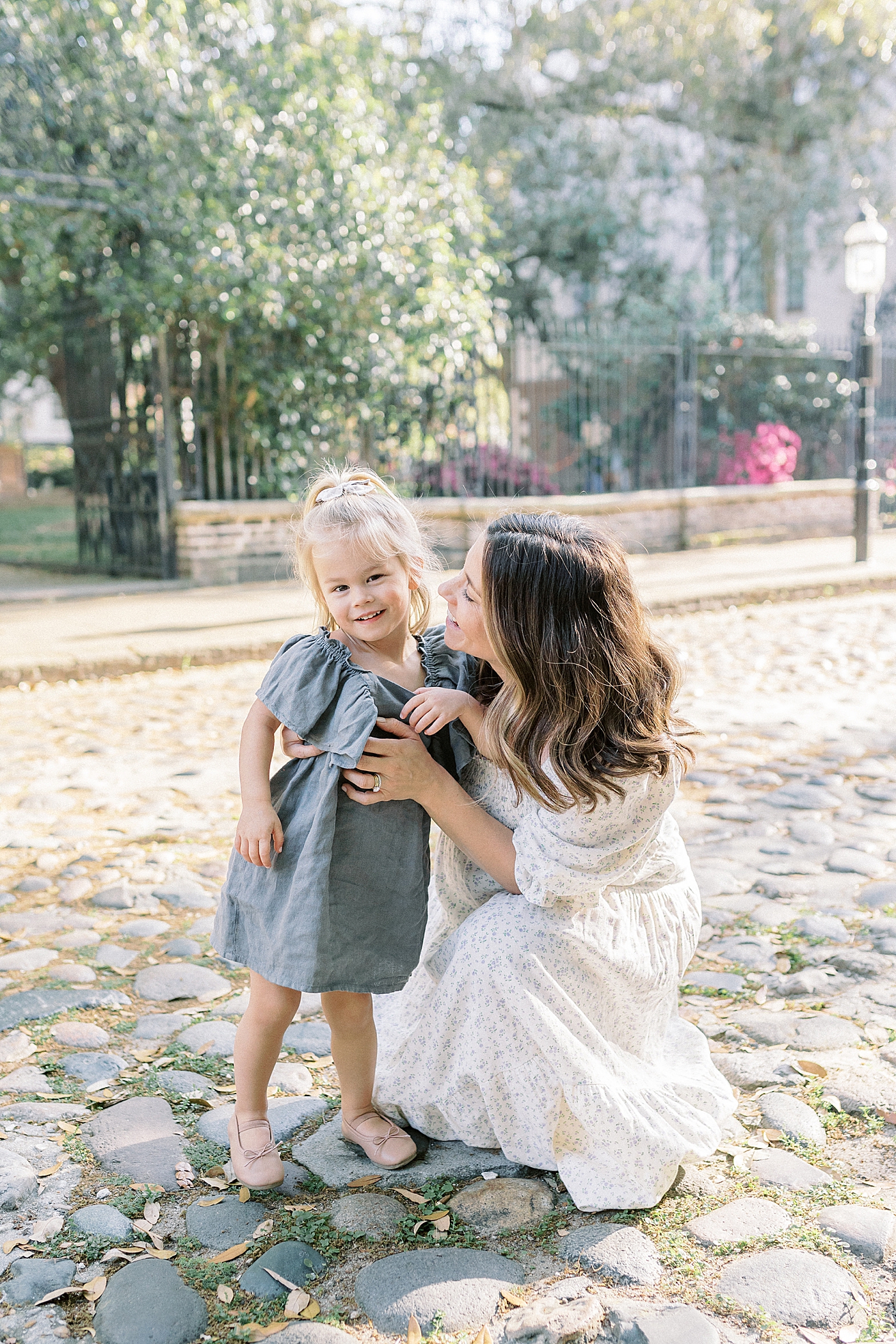 Expecting mother kneeling down beside her daughter on a cobblestone street during Spring Motherhood Minis in Charleston | Image by Caitlyn Motycka