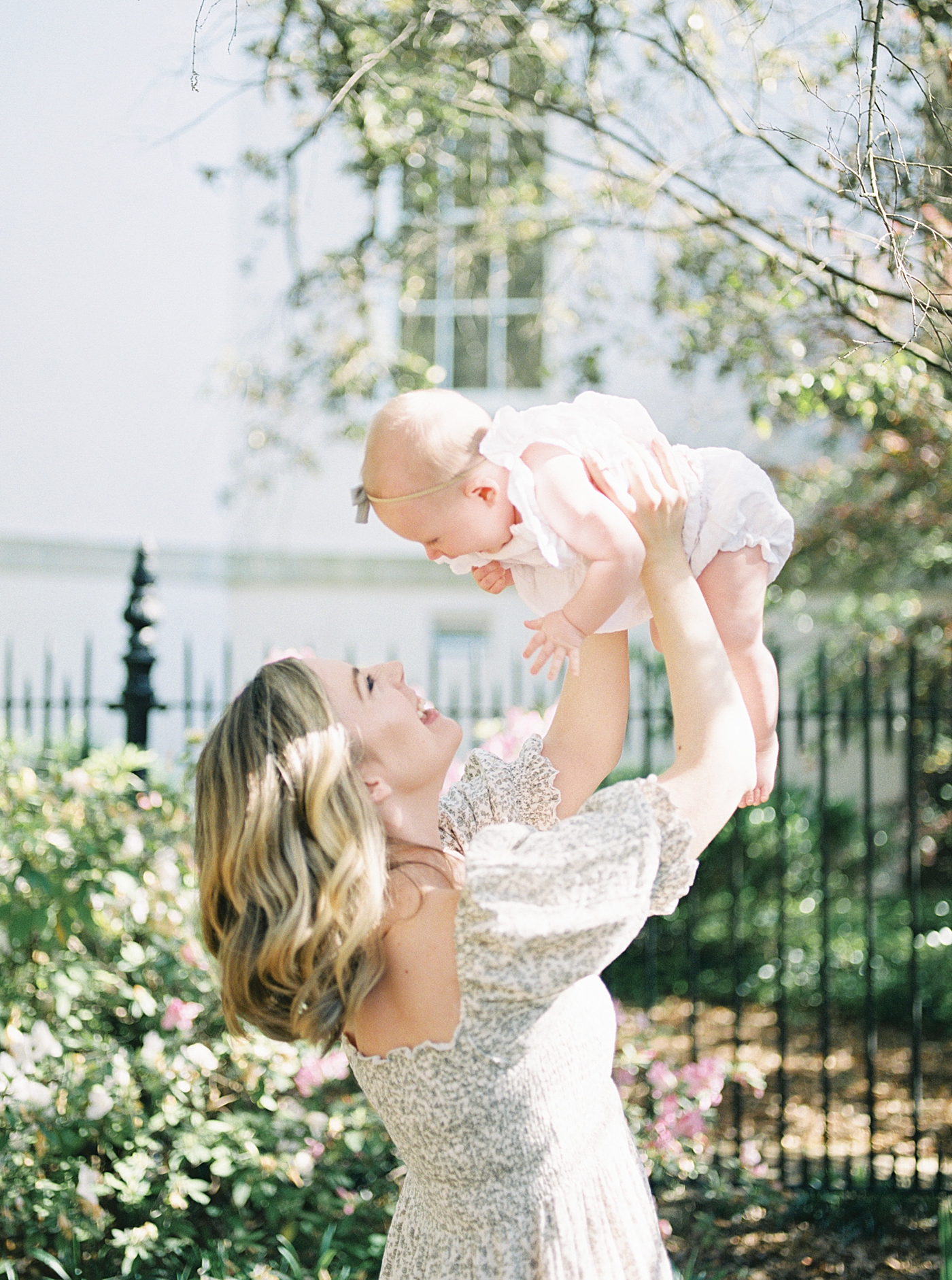 Mother in a spring dress smiling and holding her baby in the air during Spring Motherhood Minis in Charleston | Image by Caitlyn Motycka