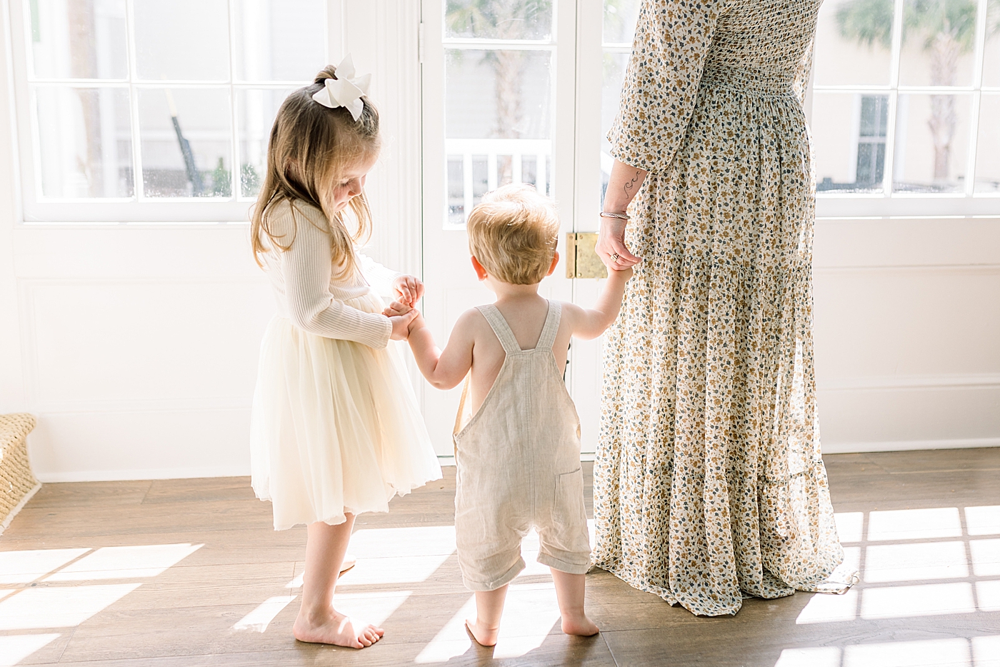Mother holding hands with her two children next to a window with sun streaming in | Image by Caitlyn Motycka 