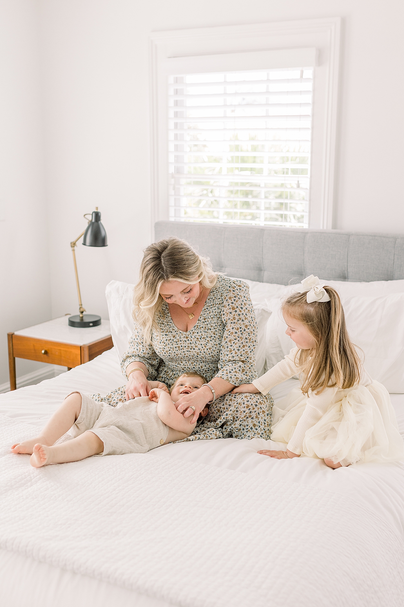 Mother in a sunny bedroom on the bed smiling, playing with her two children | Image by Caitlyn Motycka 