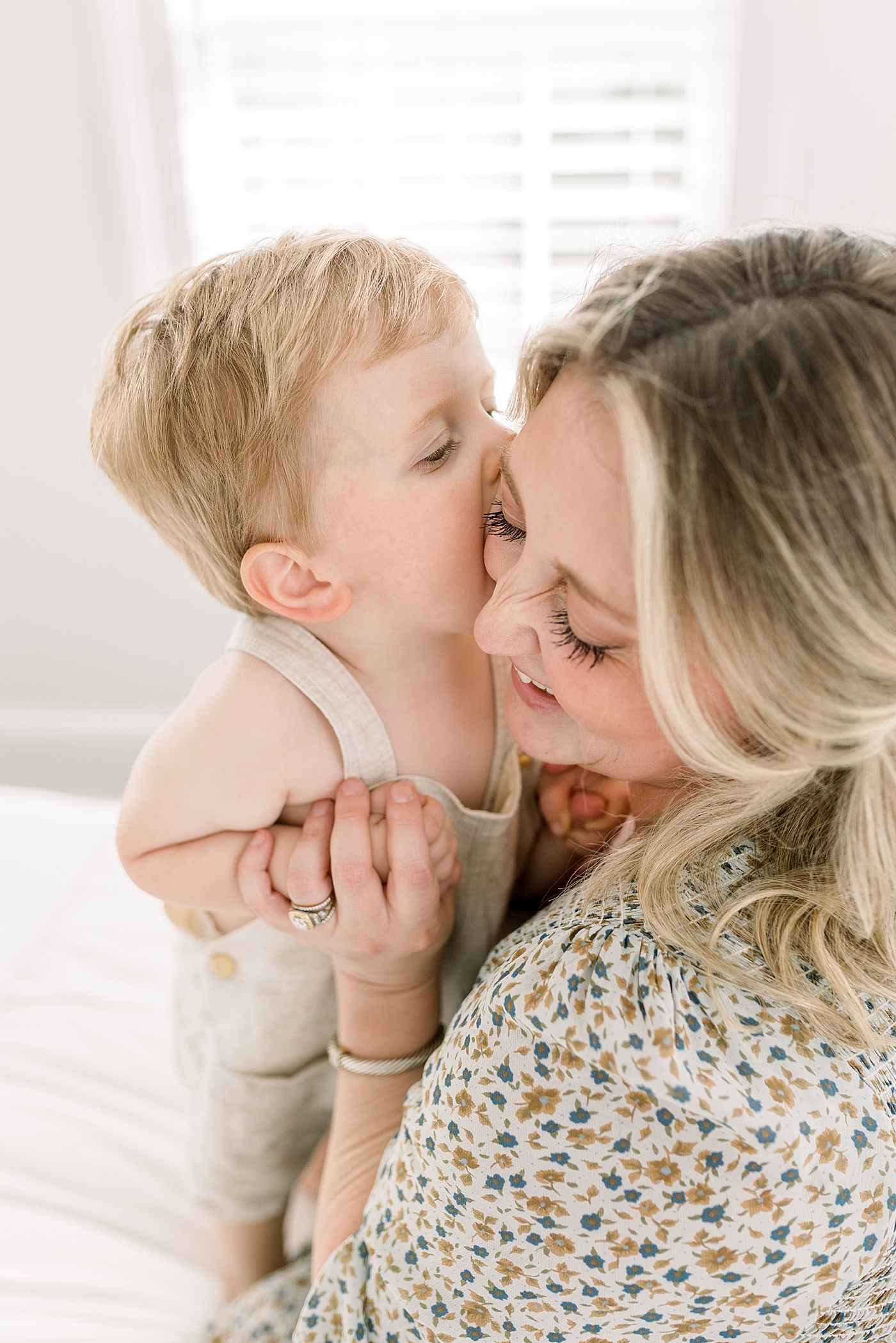 Close up of a little boy kissing his mother on the cheek while she's smiling in a brightly lit bedroom | Image by Caitlyn Motycka 