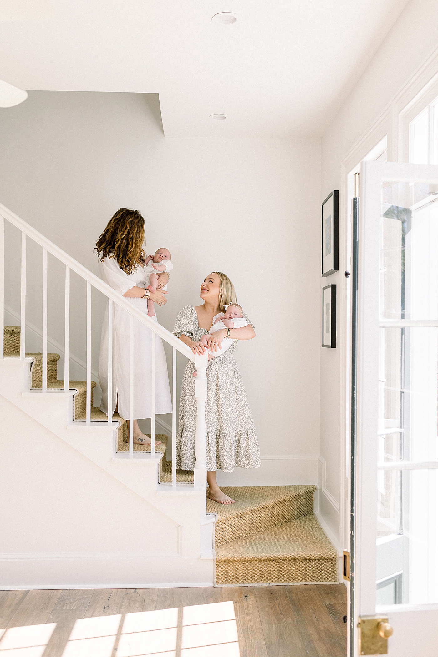 Mother and grandmother in spring dresses holding twins while walking down stairs to a simple foyer | Image by Caitlyn Motycka 