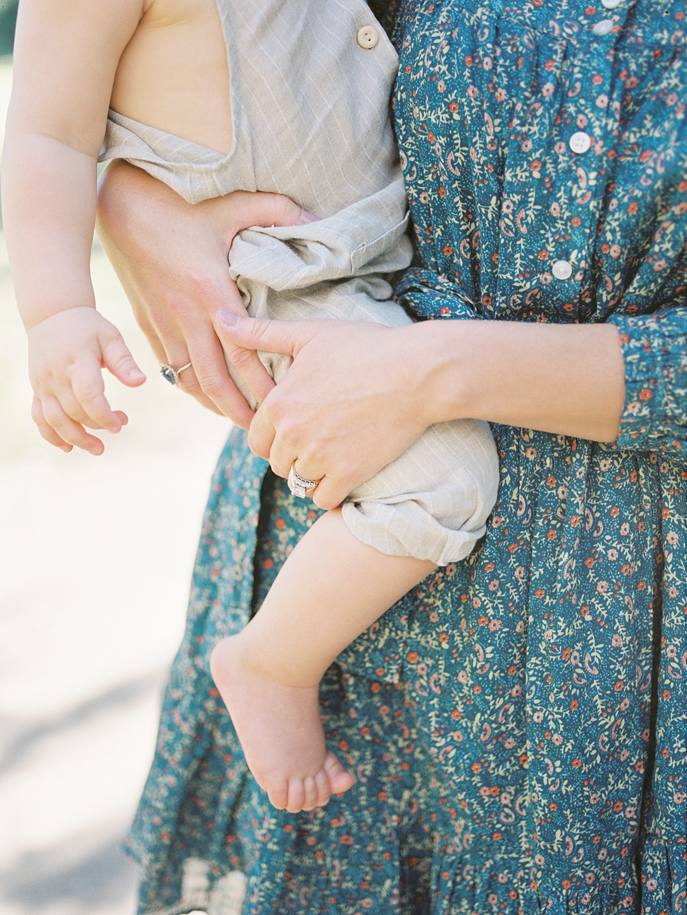 Detail of mom in floral dress holding her baby in overalls | Image by Caitlyn Motycka